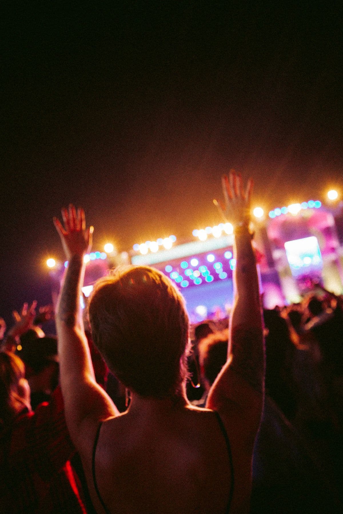 A crowd with hands raised before a lit festival stage at night